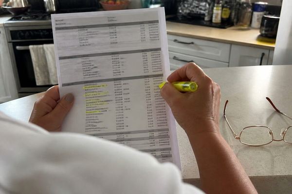 ידיים מחזיקות דף פירוט בנק מודפס על שיש המטבח, מרקר מסמן שורות קטנות ומשקפי קריאה מונחים בצד
