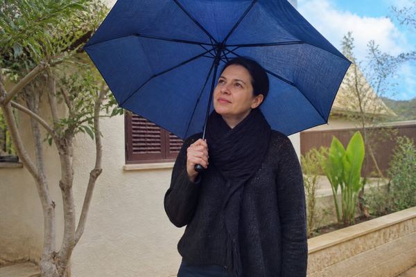 אדם בשנות הארבעים עומד מחוץ לביתו ומחזיק מטרייה פתוחה ביום שמשי חלקית, בהבעה רגועה