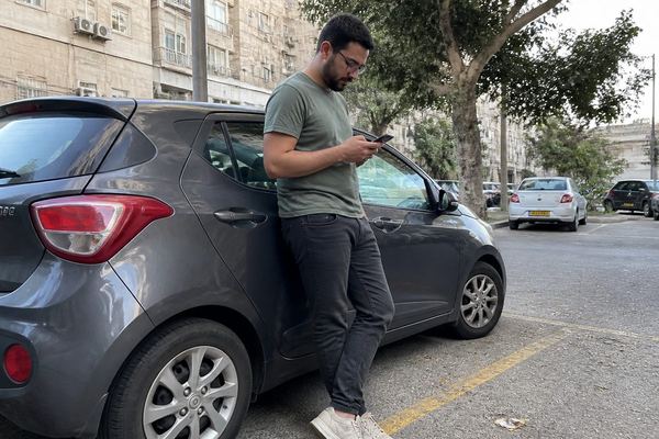 גבר בשנות ה-30 עומד ליד רכב רגיל בחניון ובודק את הטלפון, רחוב ישראלי יומיומי באור יום