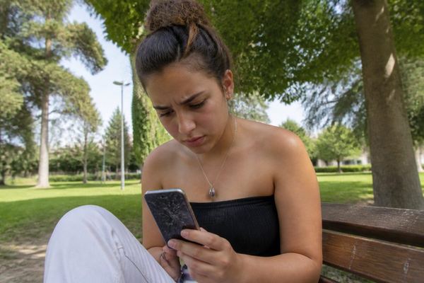צעיר יושב על ספסל בפארק ובוחן מסך טלפון סדוק בהבעה מודאגת, באור יום