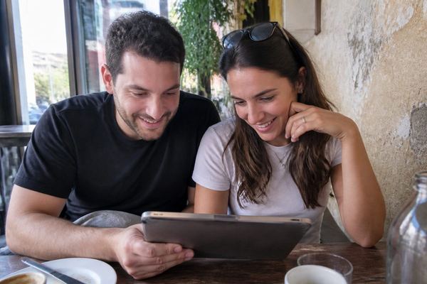זוג מאורס יושב ליד שולחן בבית קפה ומסתכל על רשימת משימות בטאבלט, מחייך, באור חלון טבעי