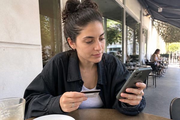 צעיר מחזיק כרטיס אשראי וטלפון ליד שולחן בבית קפה ישראלי, בהבעה מהורהרת