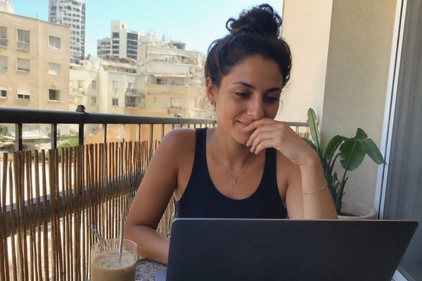 אישה בשנות ה-30 יושבת במרפסת שטופת שמש בתל אביב עם מחשב נייד וקפה קר, ובודקת את חשבון הבנק בחיוך מהורהר