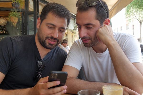 שני חברים בשנות ה-30 יושבים בבית קפה ומסתכלים יחד על תמונות של דירות בטלפון, באור טבעי