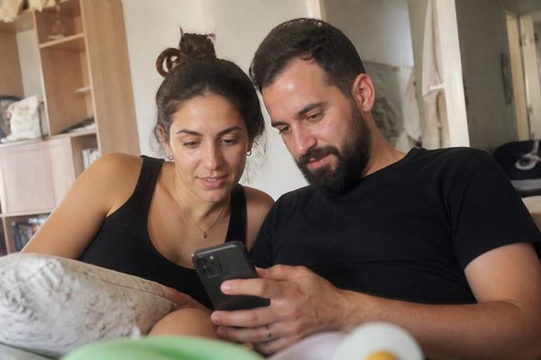 הורים טריים בשנות ה-30 בבית, כמה פריטי תינוקות סביבם, מסתכלים על רשימה בטלפון באור חם
