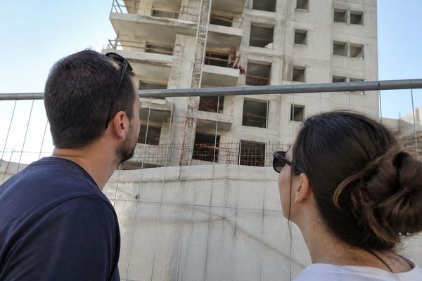 זוג בשנות ה-30 מביט מבעד לגדר בבניין מגורים בבנייה, בהבעה מלאת תקווה, באור יום