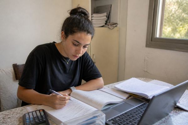 סטודנט צעיר יושב ליד שולחן עם ספרי לימוד, מחשב נייד ומחשבון, מרוכז, בחדר צנוע באור יום