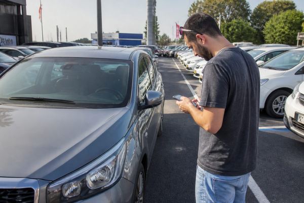 אדם עומד במגרש של סוכנות רכב באור יום, מביט ברכב רגיל ובודק משהו בטלפון