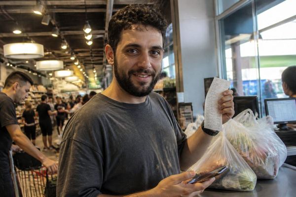 גבר בסוף שנות ה-20 בקופה בסופרמרקט, עייף אך מחייך, מחזיק קבלה וטלפון ליד שקיות קניות על הדלפק