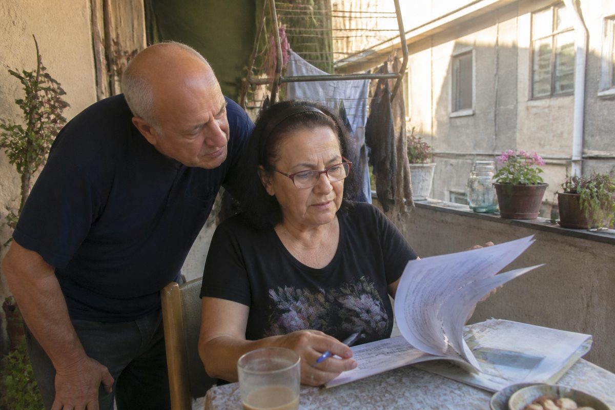 זוג מבוגר במרפסת ירושלמית עוברים יחד על דוח פנסיה מודפס, כוס קפה על השולחן