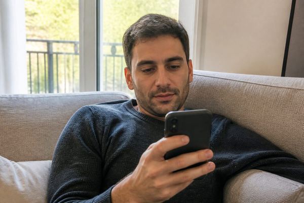 אדם יושב על ספה באור יום רך וקורא חדשות כלכליות בטלפון, בהבעה רגועה