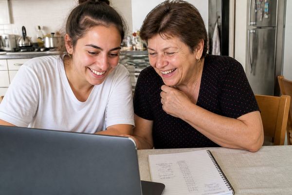 סבתא ונכדתה הבוגרת צוחקות יחד מול מחשב נייד ליד שולחן מטבח, ולצידן מחברת עם מספרים בכתב יד