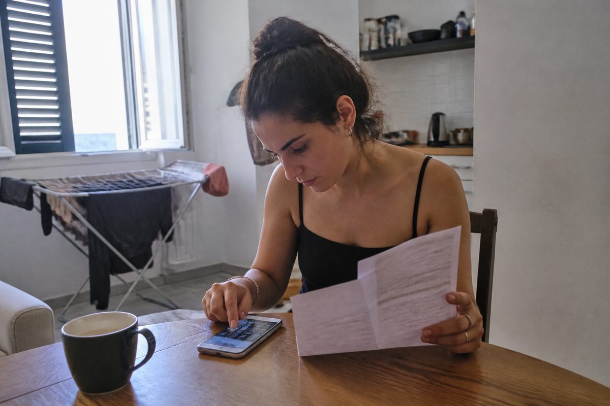 צעירה יושבת ליד שולחן מטבח קטן, מחזיקה תלוש שכר מודפס ומחשבת בטלפון, לצד כוס קפה