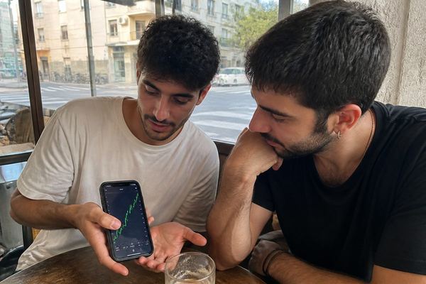 שני חברים בשנות ה-20 בבית קפה בתל אביב, אחד מראה לשני גרף מניות בטלפון, רחוב נראה מבעד לחלון