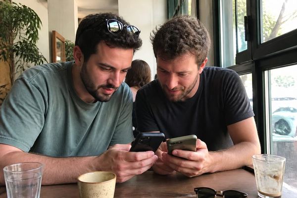 שני חברים בשנות ה-30 בבית קפה משווים משהו על שני הטלפונים שלהם זה לצד זה, אור טבעי מהחלון